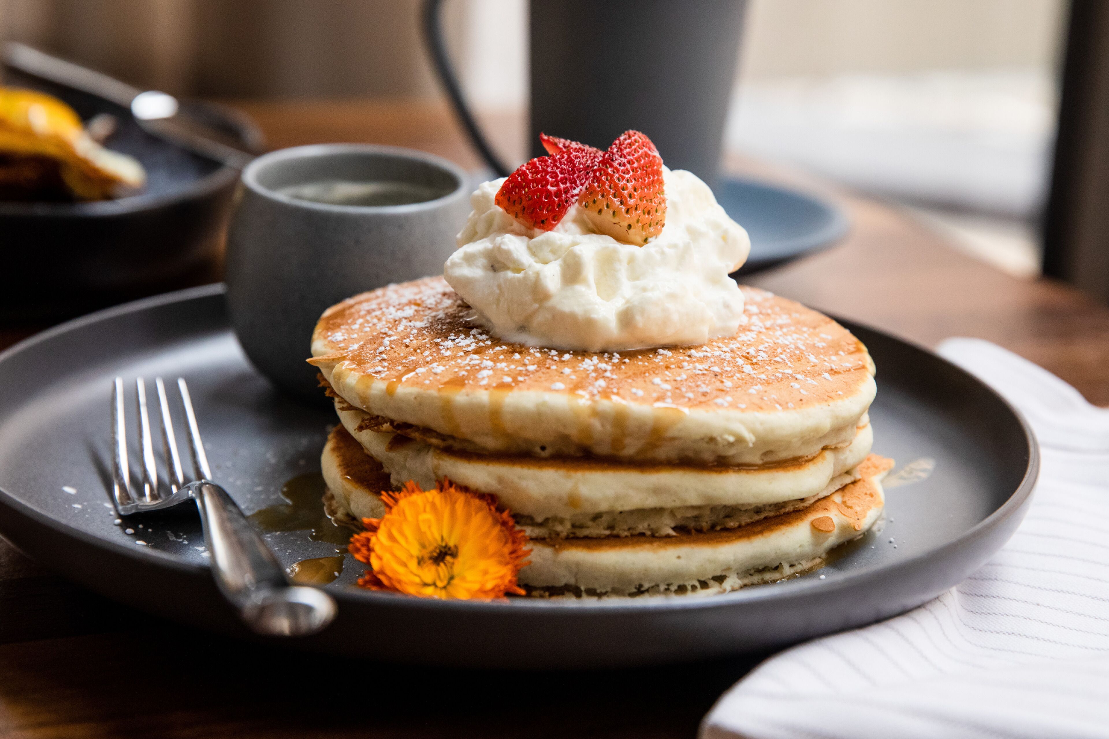 Une pile de crêpes garnie de crème fouettée et surmontée d’une fraise, le tout servi sur une assiette à côté de laquelle se trouve une fleur.