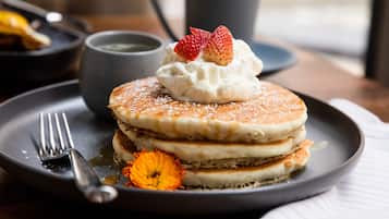 Une pile de crêpes garnie de crème fouettée et surmontée d’une fraise, le tout servi sur une assiette à côté de laquelle se trouve une fleur.