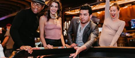A group of people standing at a craps table in a casino with a roulette wheel and chips visible.
