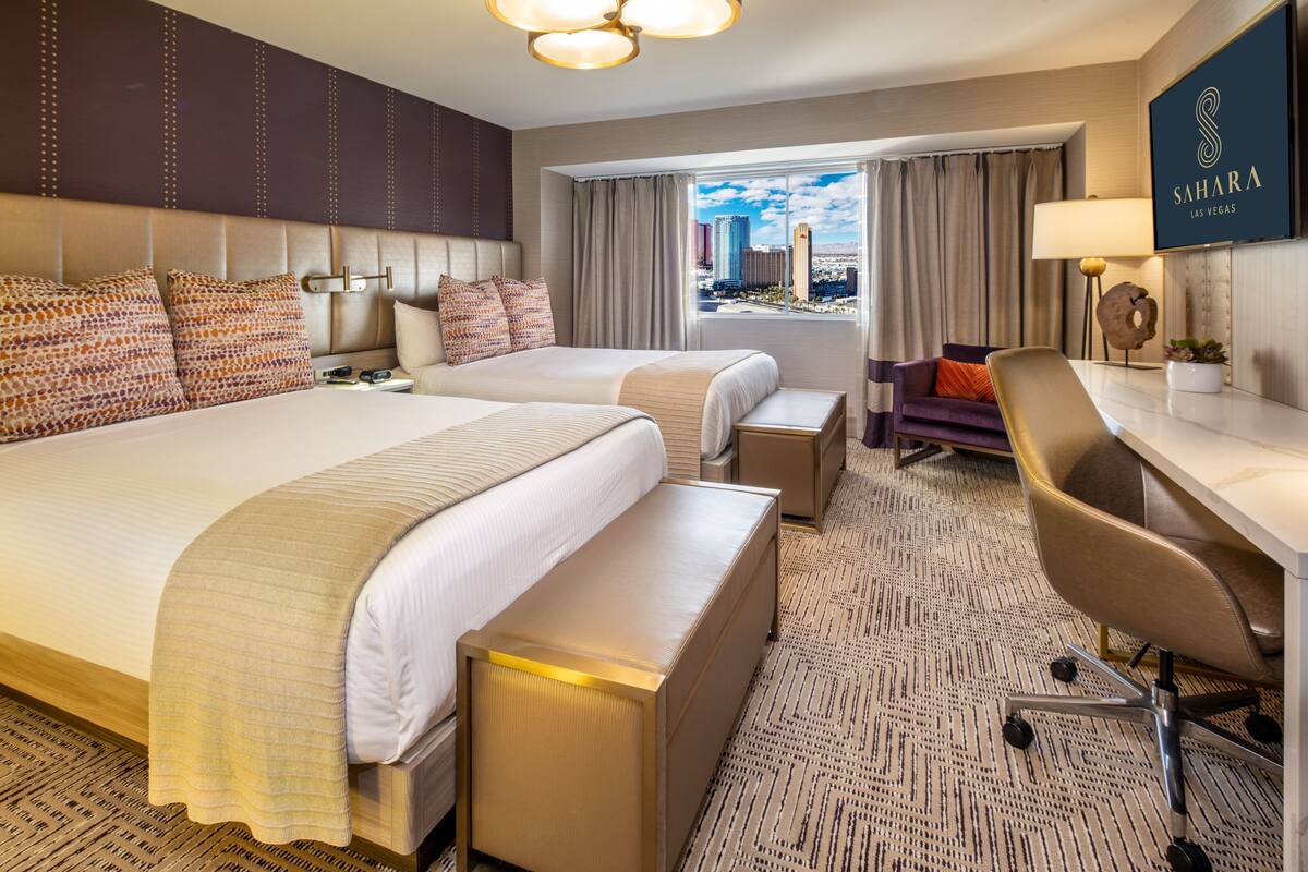 A hotel room with two beds, a desk, a chair, and a view of the city.