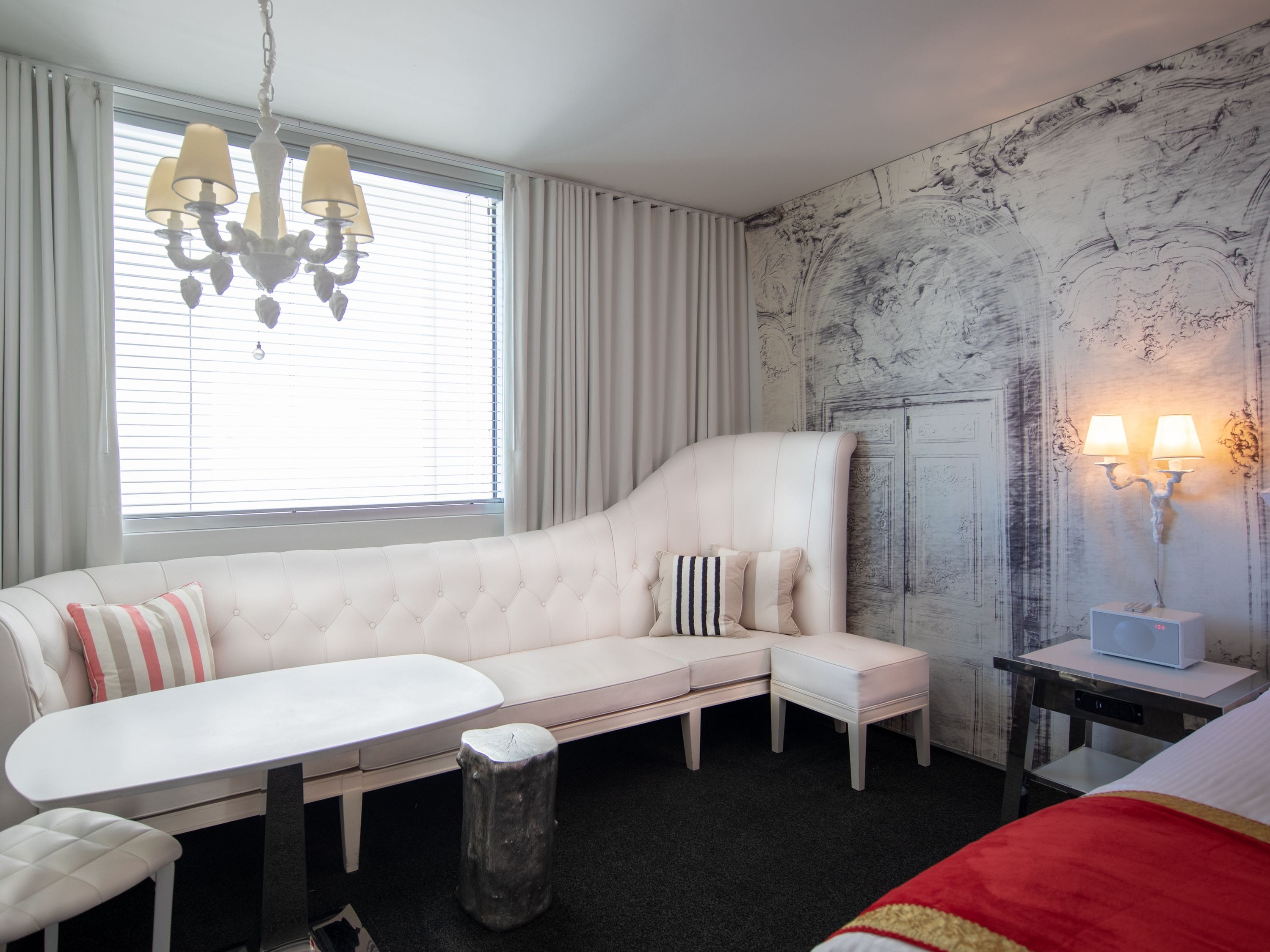 A modern hotel room with a white sofa, a small table, a bed with a red blanket, a chandelier, and a wall-mounted lamp.