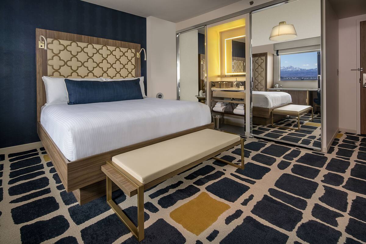 A modern hotel room with a large bed, a bench, a mirror, and a view of mountains through the window.