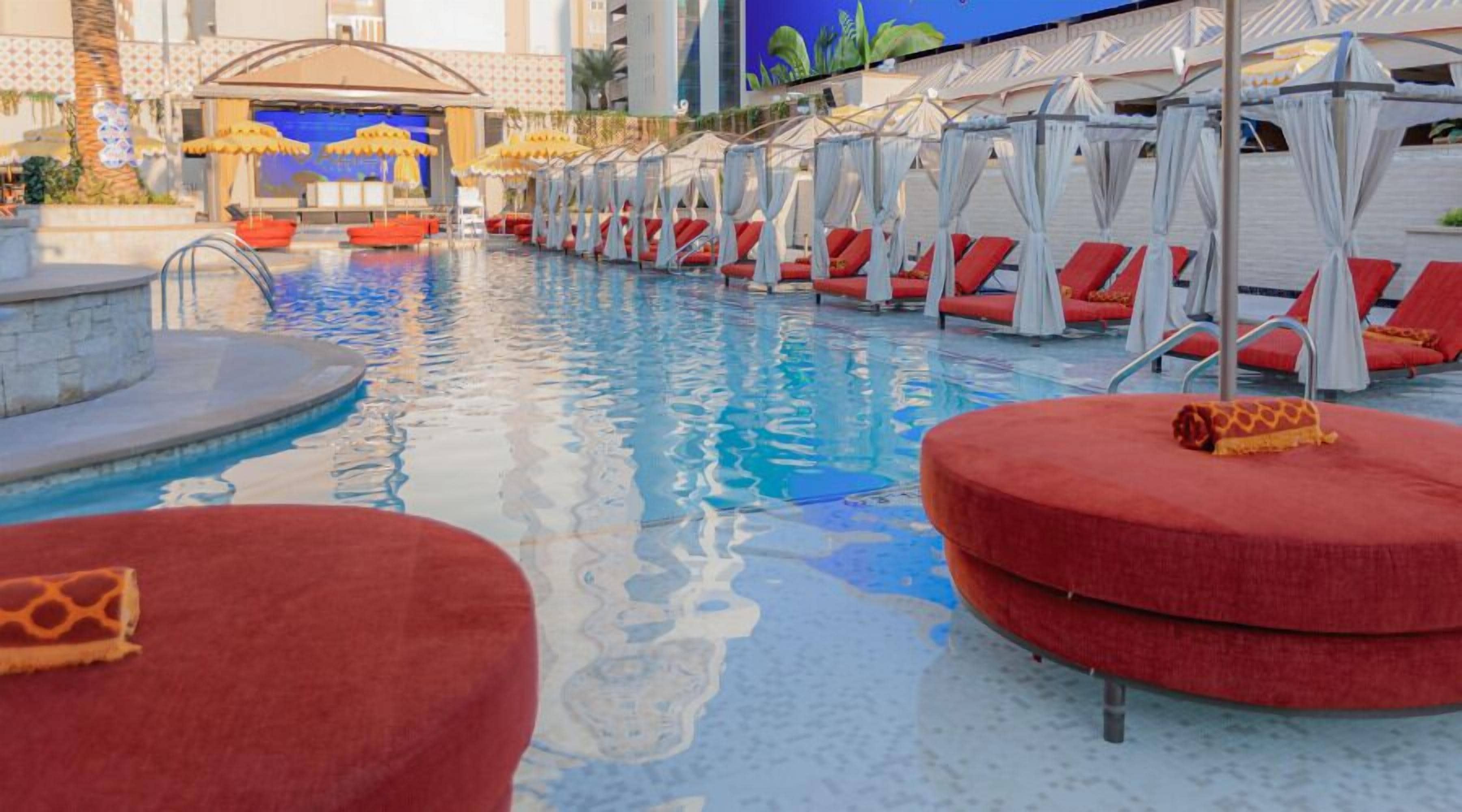 Zona de piscina con sillas de descanso rojas y toldos blancos.