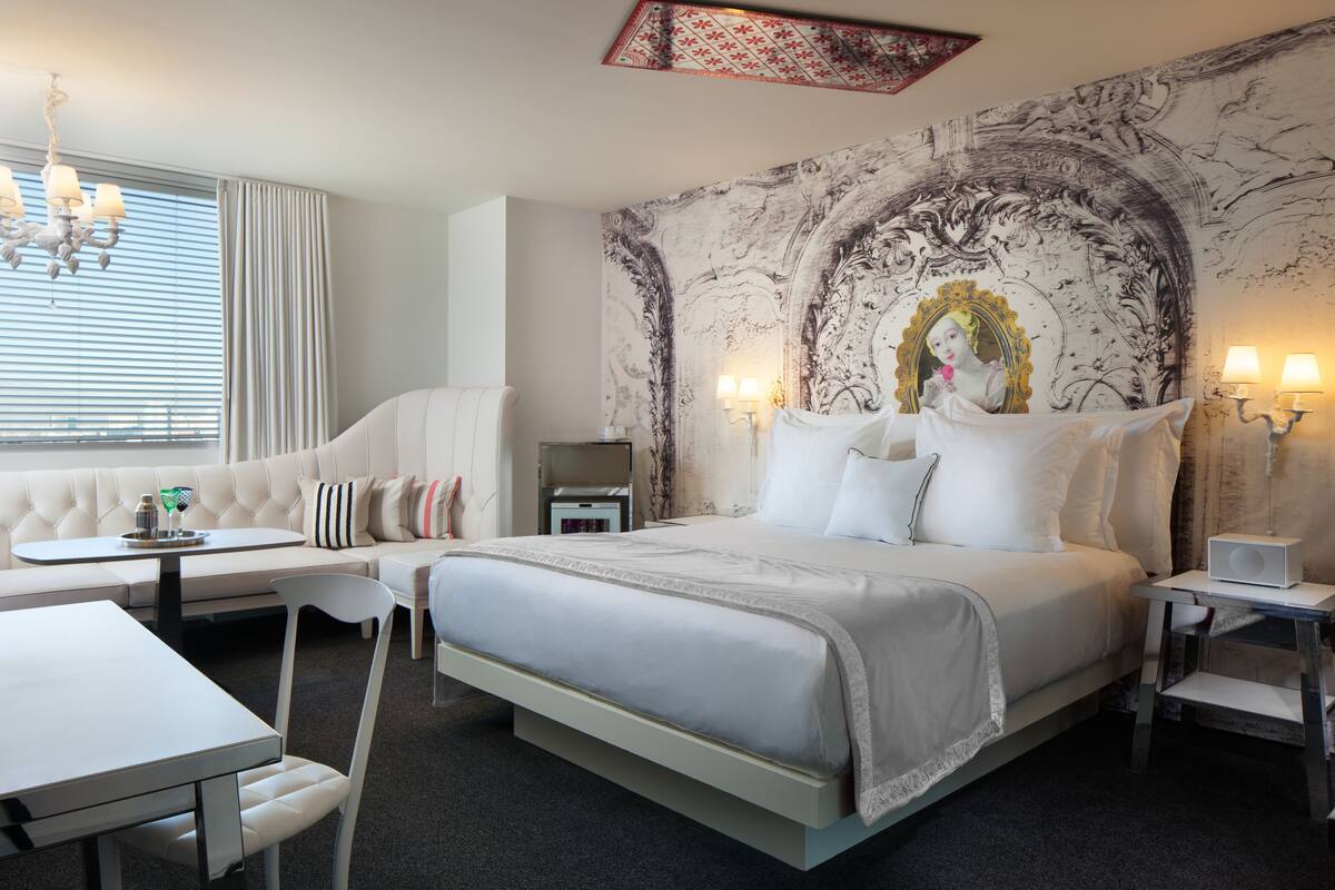 A modern hotel room with a large bed, a sofa, a dining table, and a chandelier.