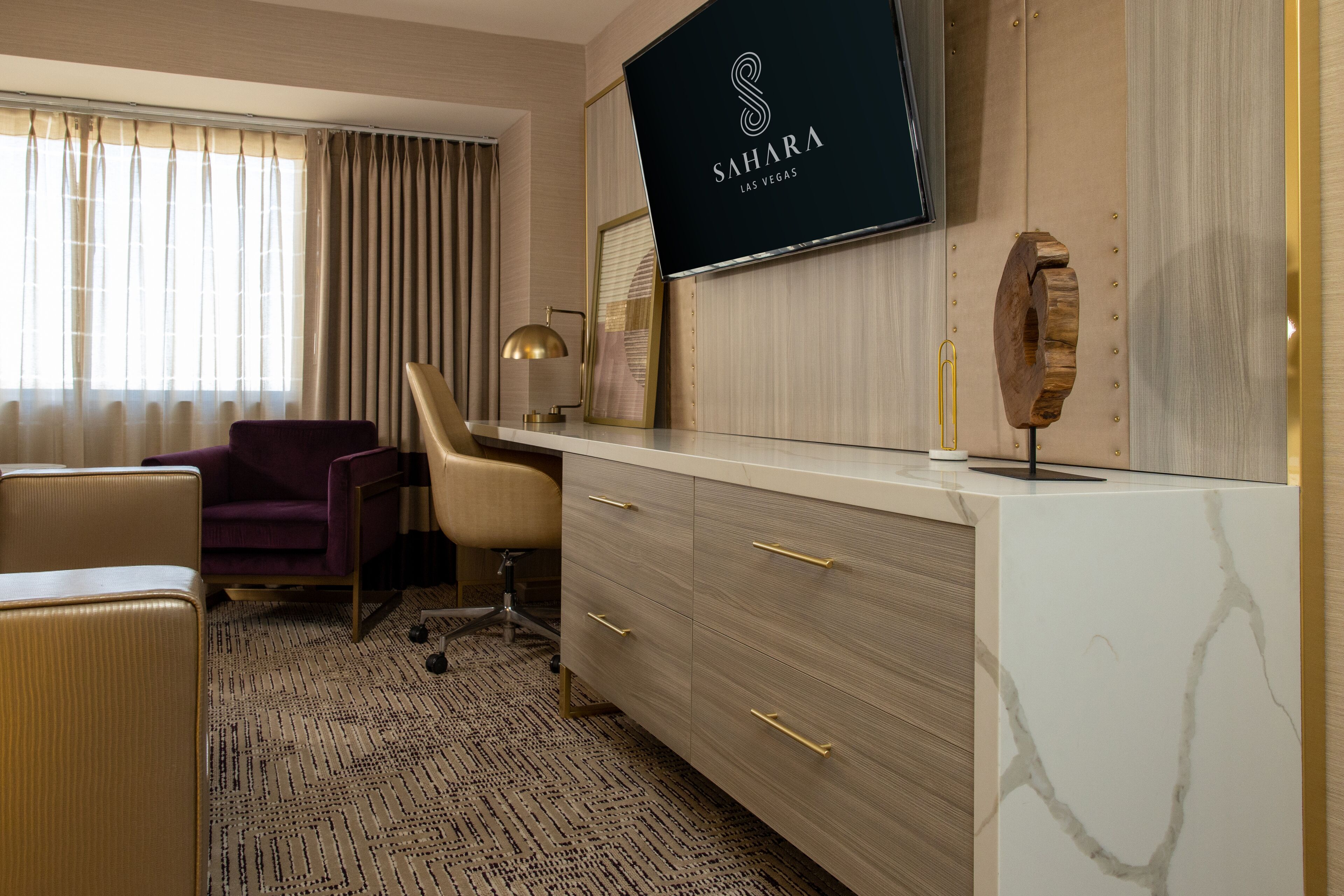 A hotel room with a large flat-screen TV mounted on the wall, a desk with drawers, a chair, and a sofa.