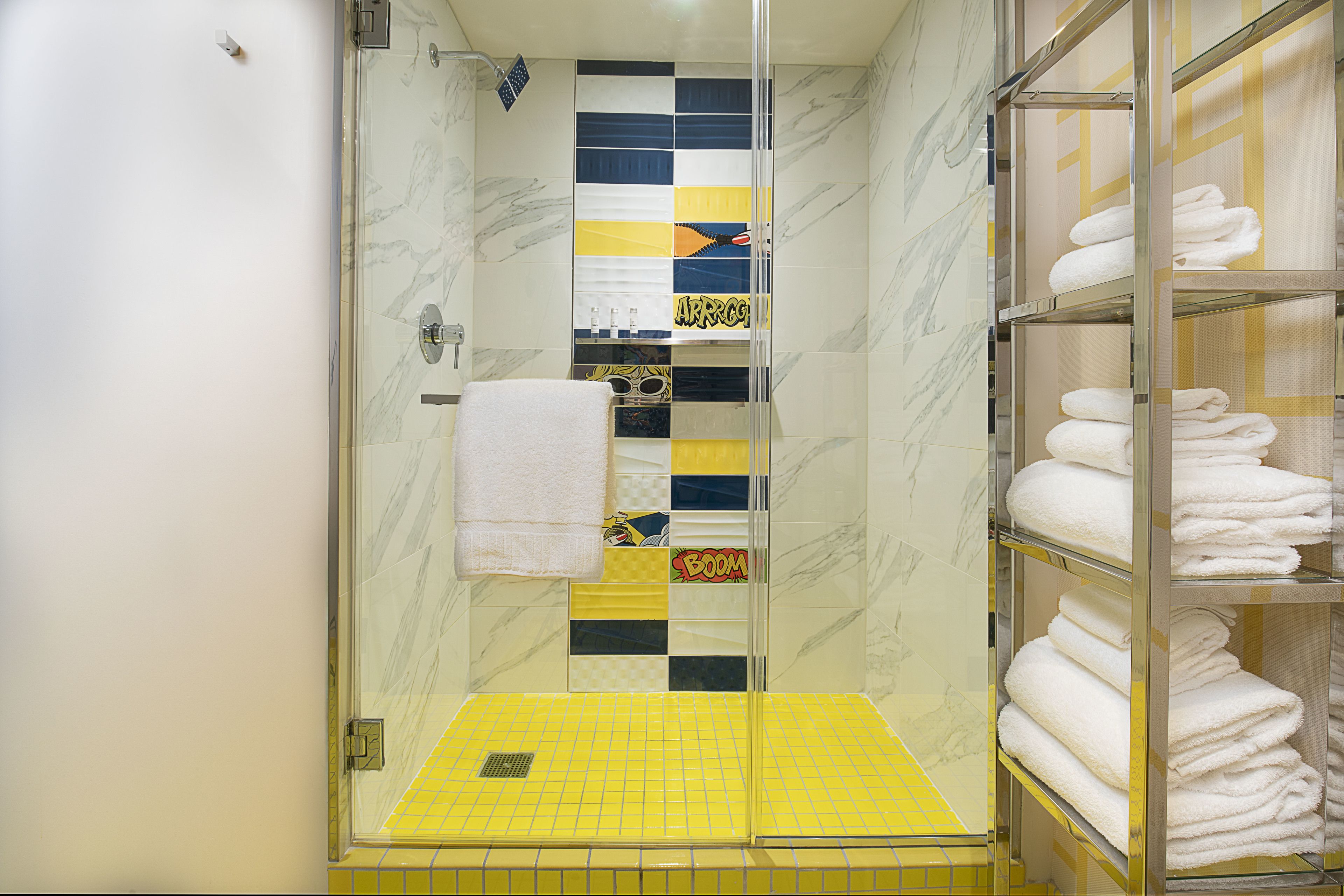 Un baño con cabina de ducha de vidrio, piso con baldosas amarillas y un estante con toallas dobladas con esmero.