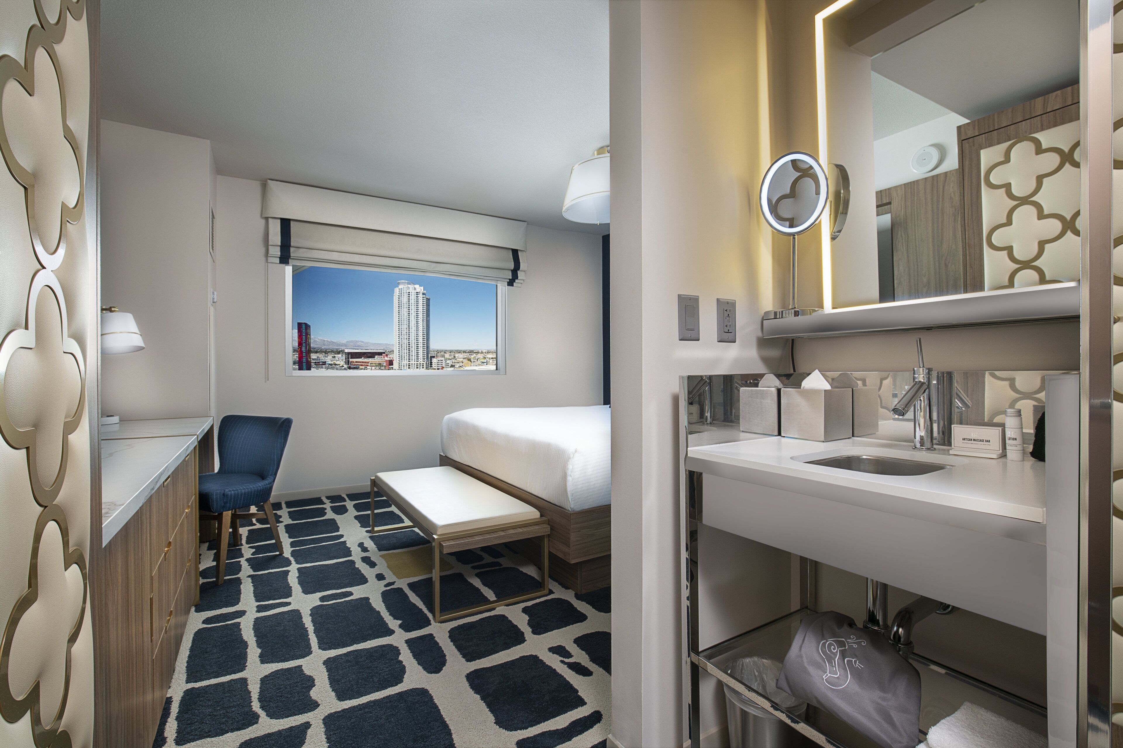 A hotel room with a bed, a desk, a chair, a small table, a bathroom with a sink, mirror, and towel rack.