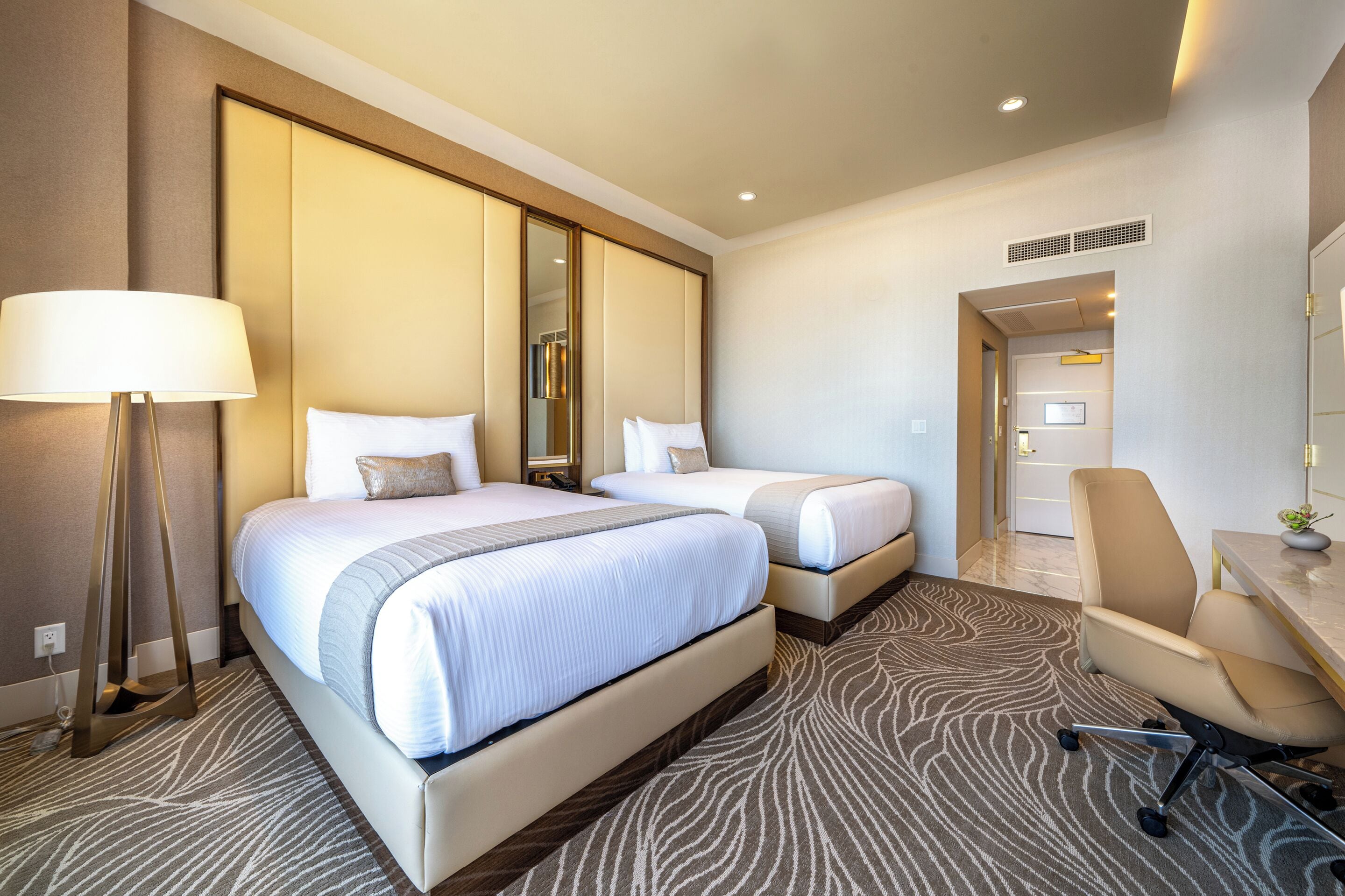Ein Hotelzimmer mit zwei Betten, einem Schreibtisch, einem Stuhl, einer Lampe und einem gemusterten Teppich.