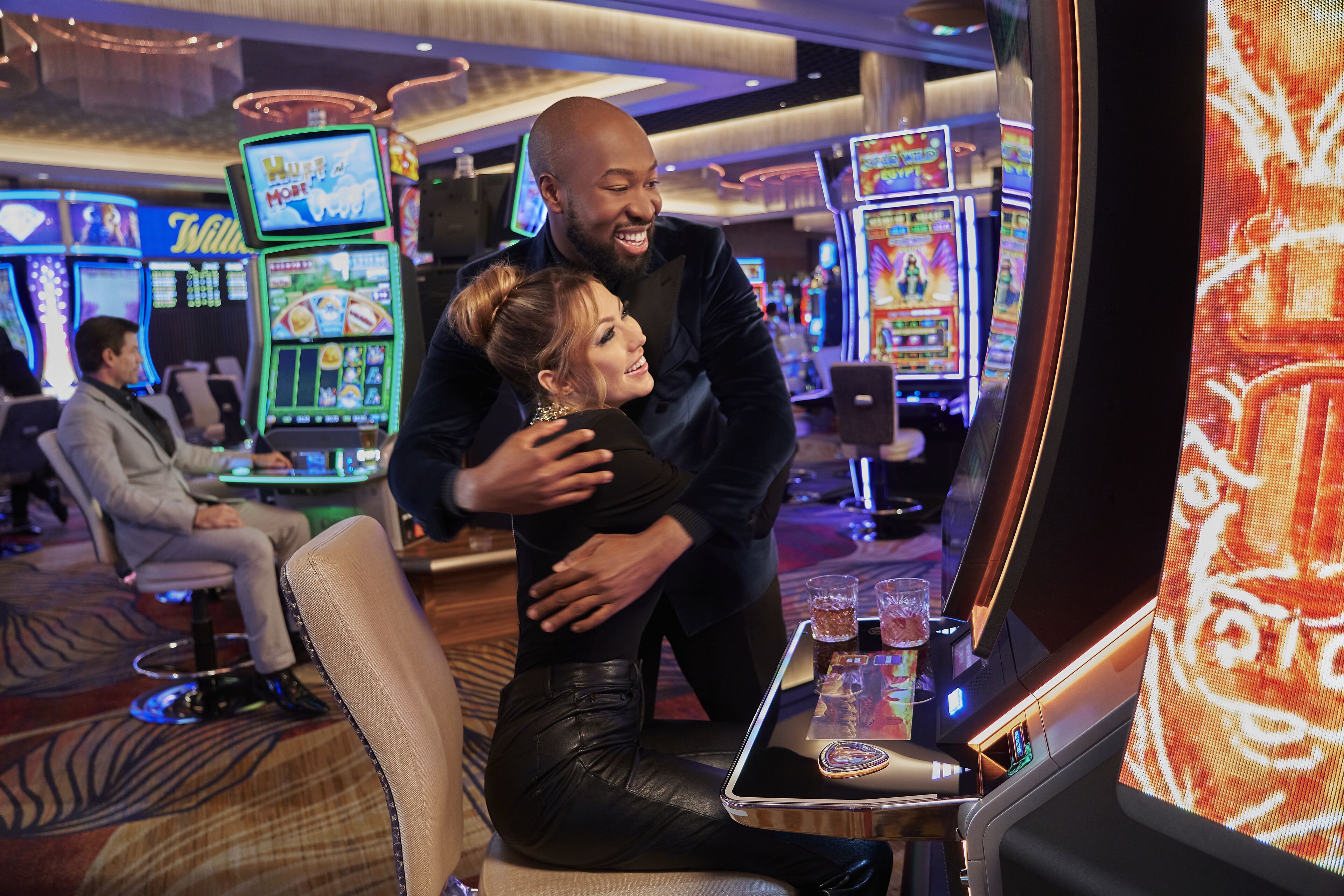Una pareja abrazándose en un casino, con máquinas tragamonedas y una pantalla grande al fondo.