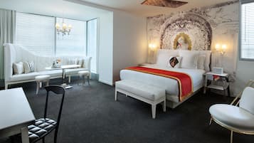 Una habitación de hotel moderna con una cama grande, una zona de estar con sofá y sillones, una mesita y un ventanal con persianas.