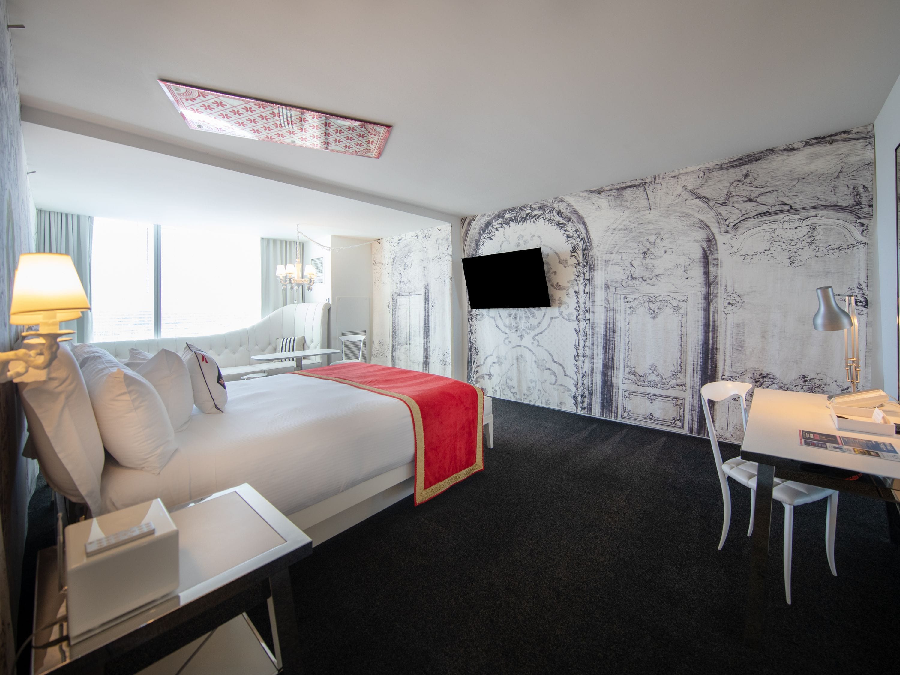 A hotel room with a large bed, a desk, a chair, a TV mounted on the wall, and a window with curtains.