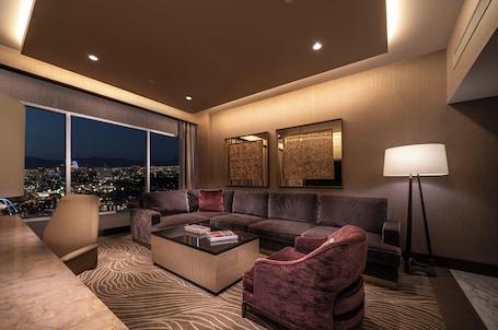 A modern living room with a large sectional sofa, a coffee table, and a floor lamp. There are two wall-mounted decorative panels and a large window offering a city view.. SAHARA Las Vegas