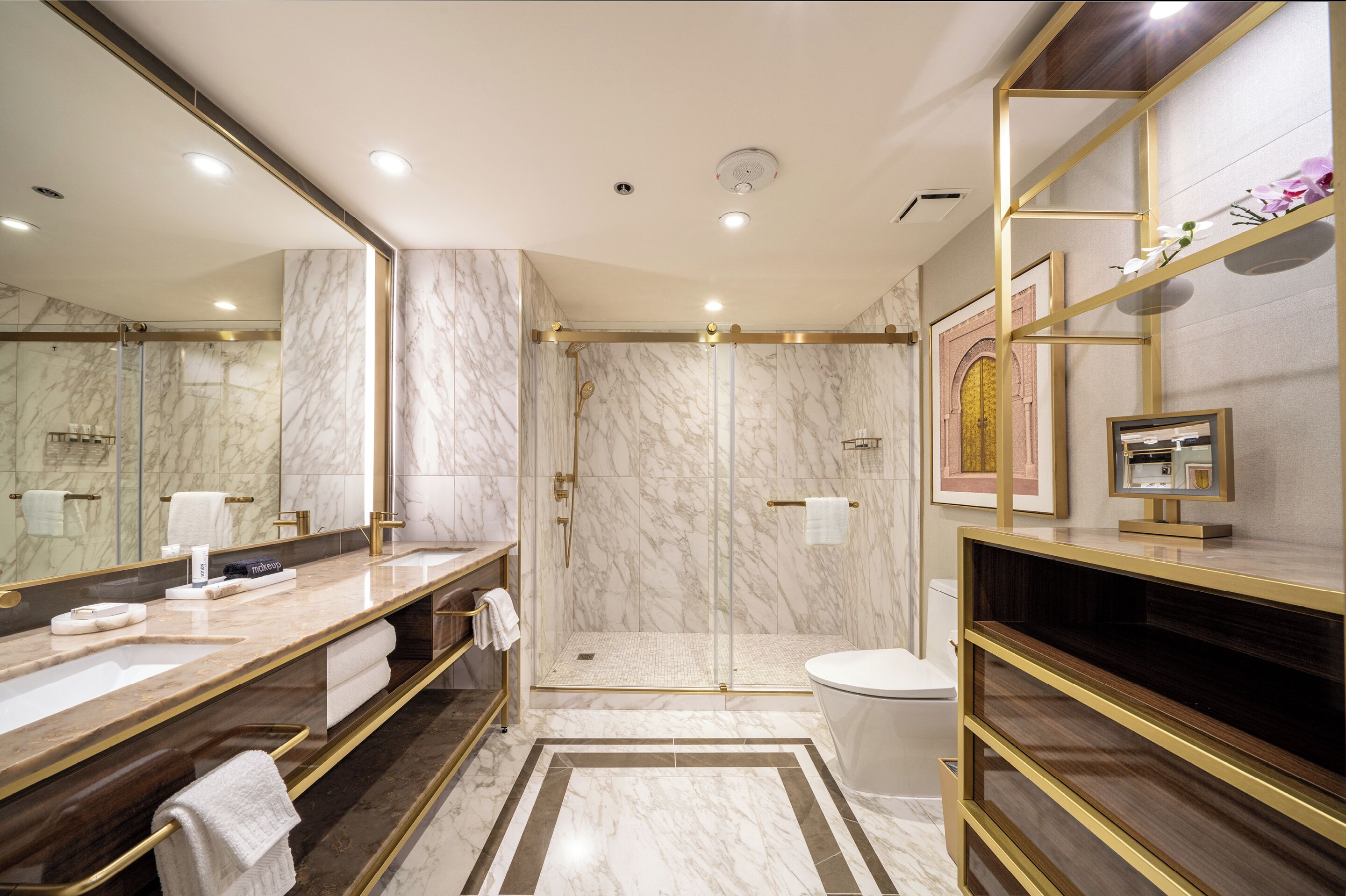 A modern bathroom with a large mirror, marble walls, a glass shower enclosure, and a vanity with a gold frame.