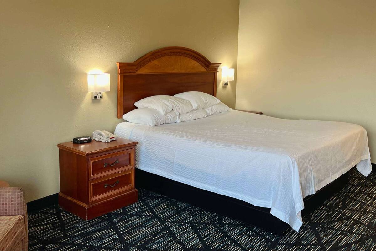 A hotel room with a wooden headboard, a white bedspread, two bedside lamps, a wooden nightstand with a phone, and a patterned carpet.