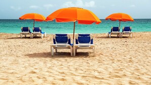 A beach with four blue lounge chairs under orange umbrellas, facing the ocean. - DoubleTree by Hilton San Juan (San Juan)