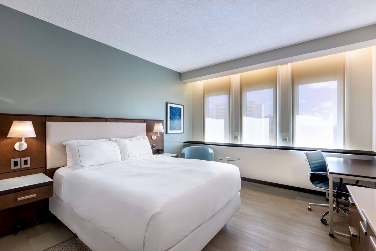 A hotel room with a large bed, bedside lamps, a desk with a chair, and a window with a city view.