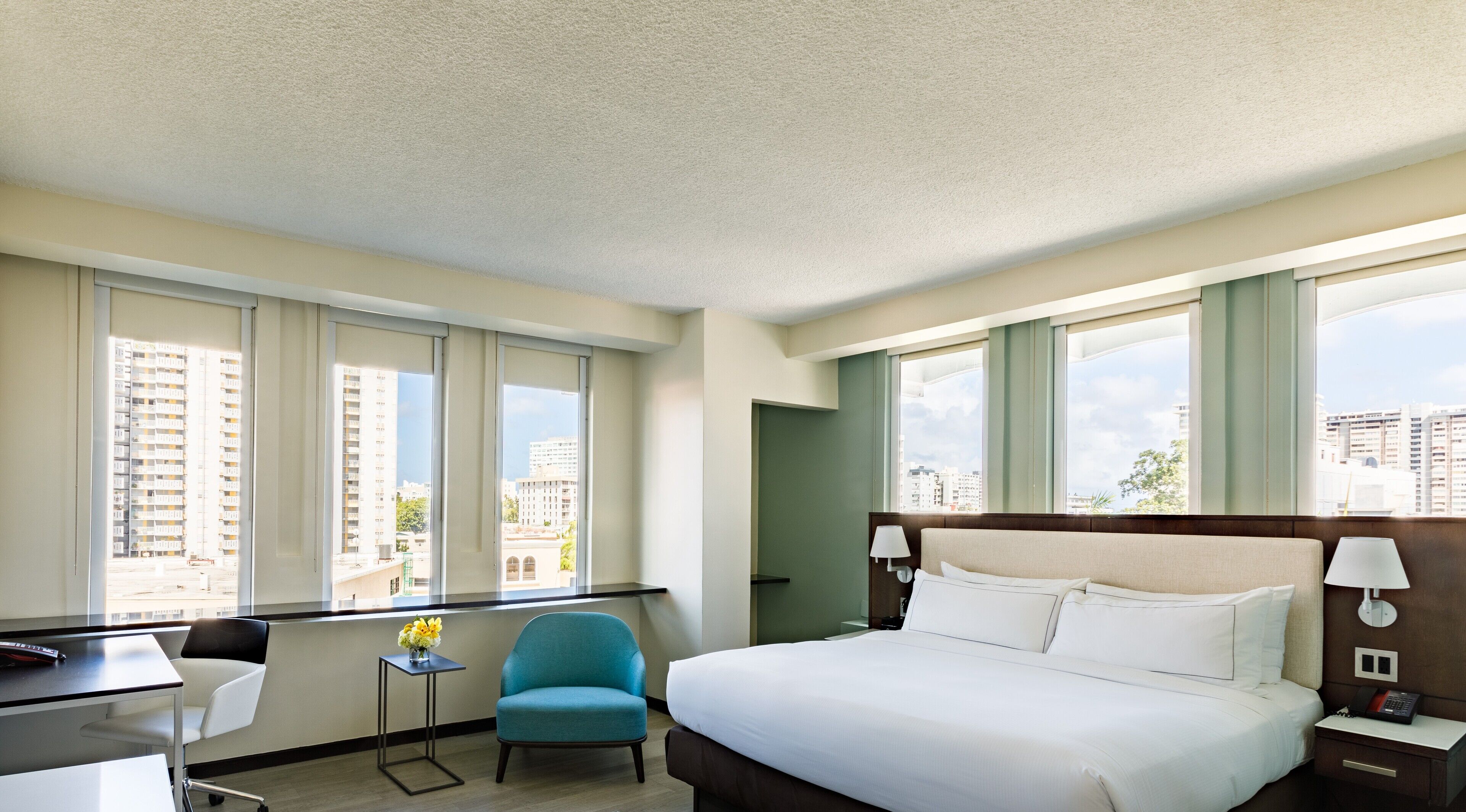 Habitación de hotel con cama, escritorio, silla y amplios ventanales con vistas a la ciudad.