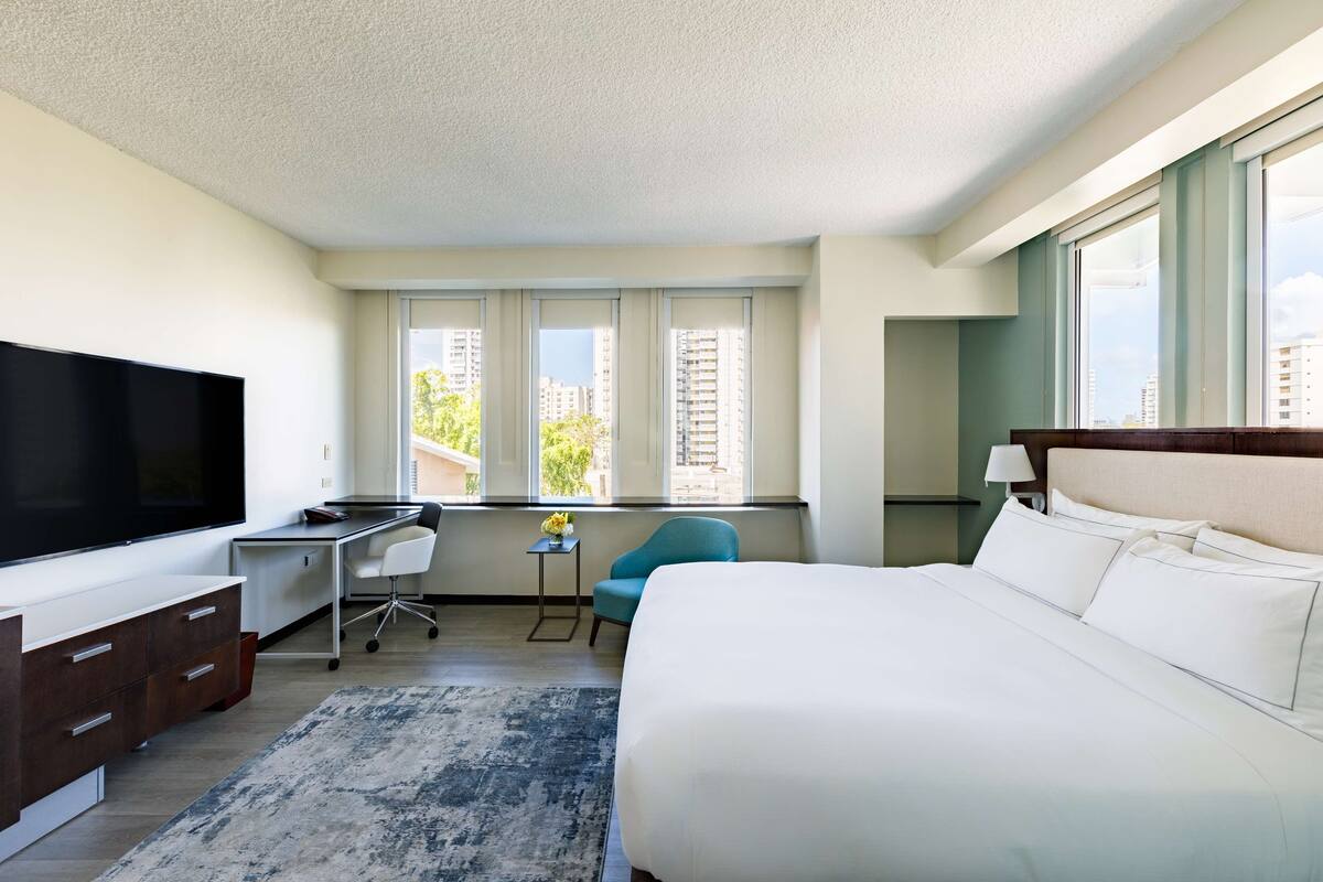 A hotel room with a large bed, a desk with a TV, a chair, and a view of buildings through the windows.