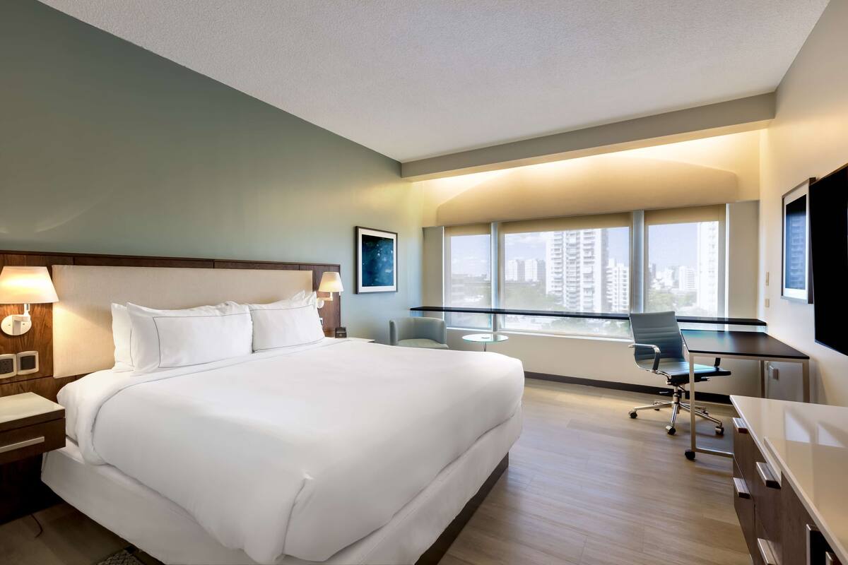 A hotel room with a large bed, a desk, a chair, and a view of the city through large windows.
