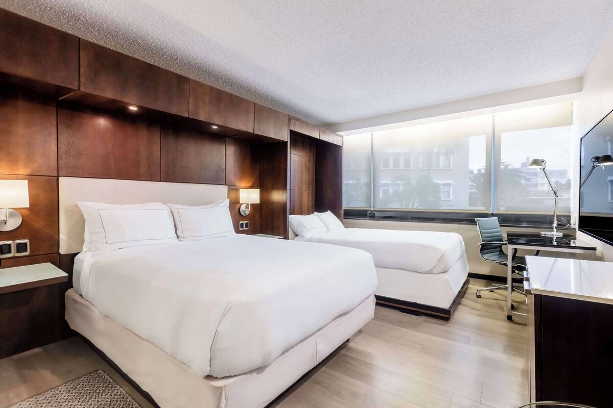 A modern hotel room with two beds, a desk, and a large window offering a city view.
