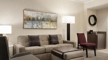 A hotel room with a sofa, armchairs, a small table, a lamp, a map artwork, a mirror, and a kitchenette.