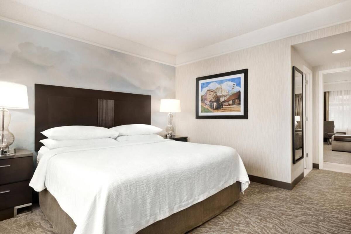 Une chambre d’hôtel avec un lit, une table de chevet, une lampe, un tableau encadré au mur et une vue sur le couloir.