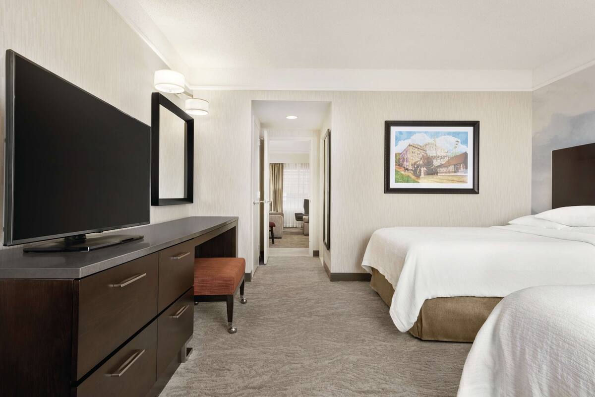 Une chambre d’hôtel équipée d’une télévision à écran plat, d’un bureau avec des tiroirs, d’un banc, de deux lits et d’un tableau encadré au mur.