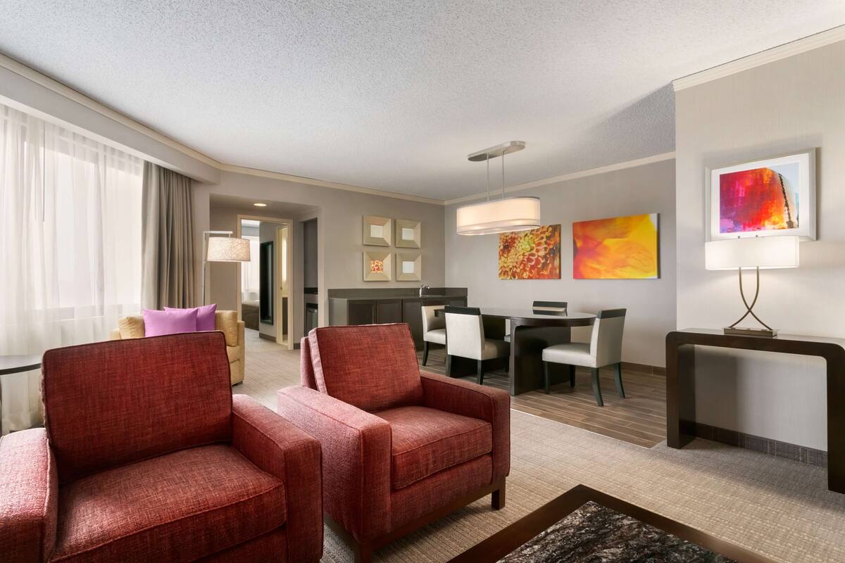 Une chambre d’hôtel avec deux fauteuils rouges, un coin repas avec une table et des chaises, et des œuvres d’art accrochées aux murs.