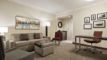A hotel room with a sofa, ottoman, a desk with a chair, a lamp, a wall with framed pictures, and a carpeted floor.