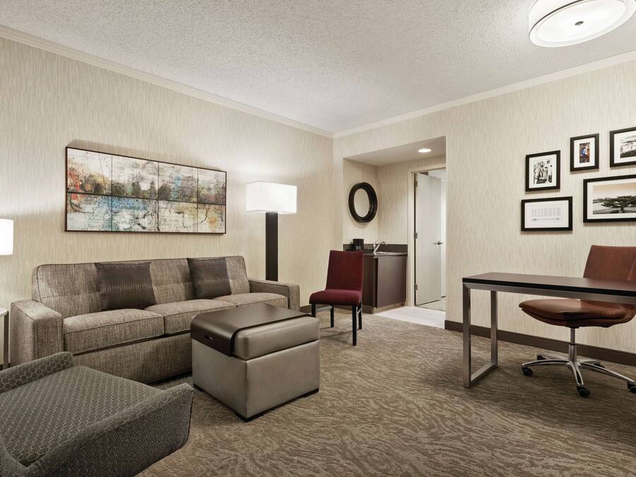 A hotel room with a sofa, ottoman, a desk with a chair, a lamp, a wall with framed pictures, and a carpeted floor.
