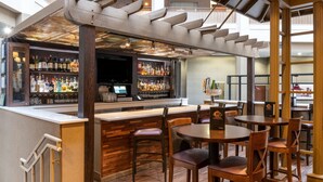 Zona de bar con sillas y mesas de madera, una barra con botellas y vasos, y una estructura de techo de madera.