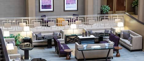 A spacious lobby with multiple seating areas, including sofas and armchairs, arranged around a central coffee table. There are two framed artworks on the wall, and a staircase is visible in the background.