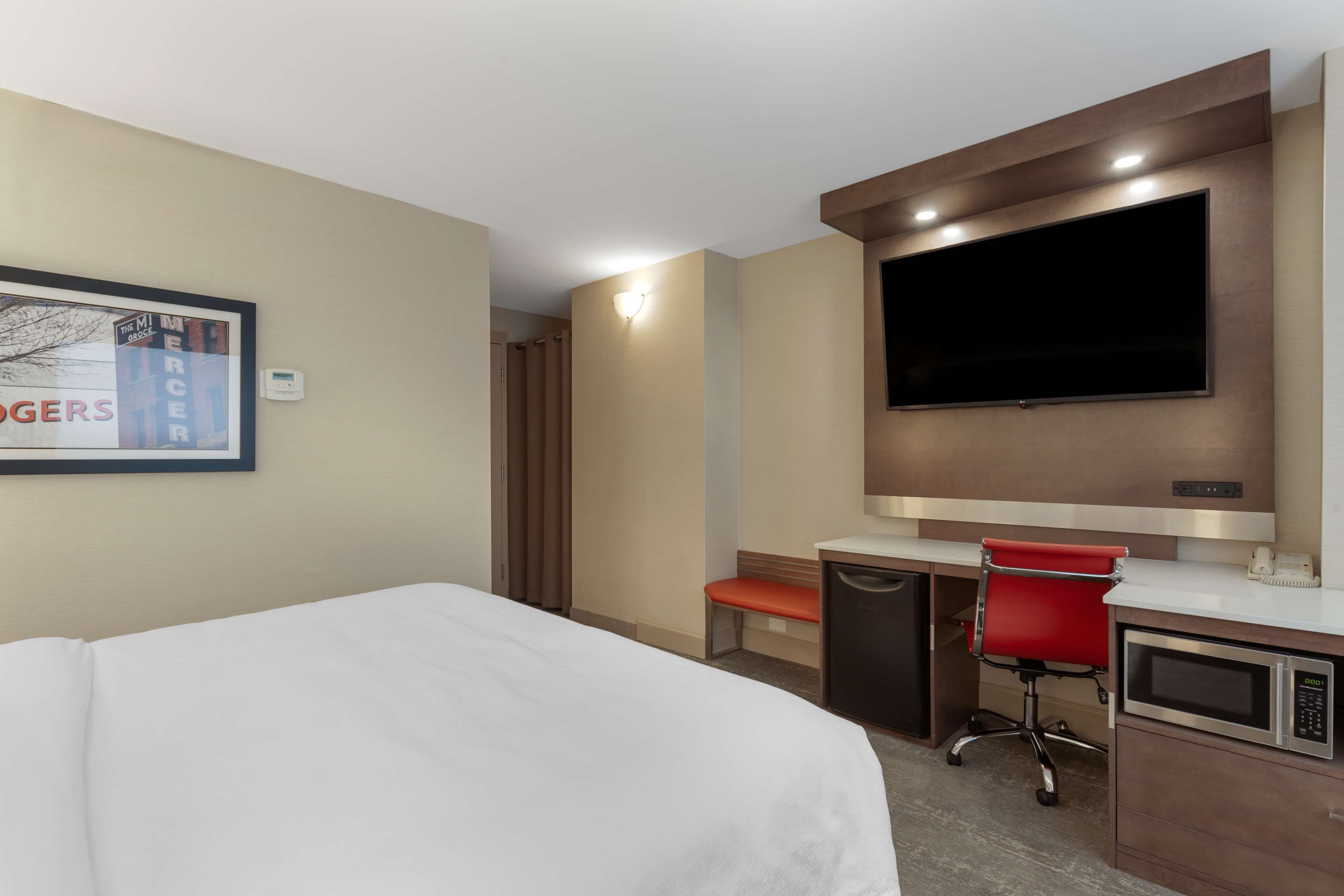 Une chambre d’hôtel comprenant un lit, un bureau, un fauteuil de bureau rouge, un téléviseur à écran plat fixé au mur et une petite kitchenette équipée d’un four à micro-ondes.