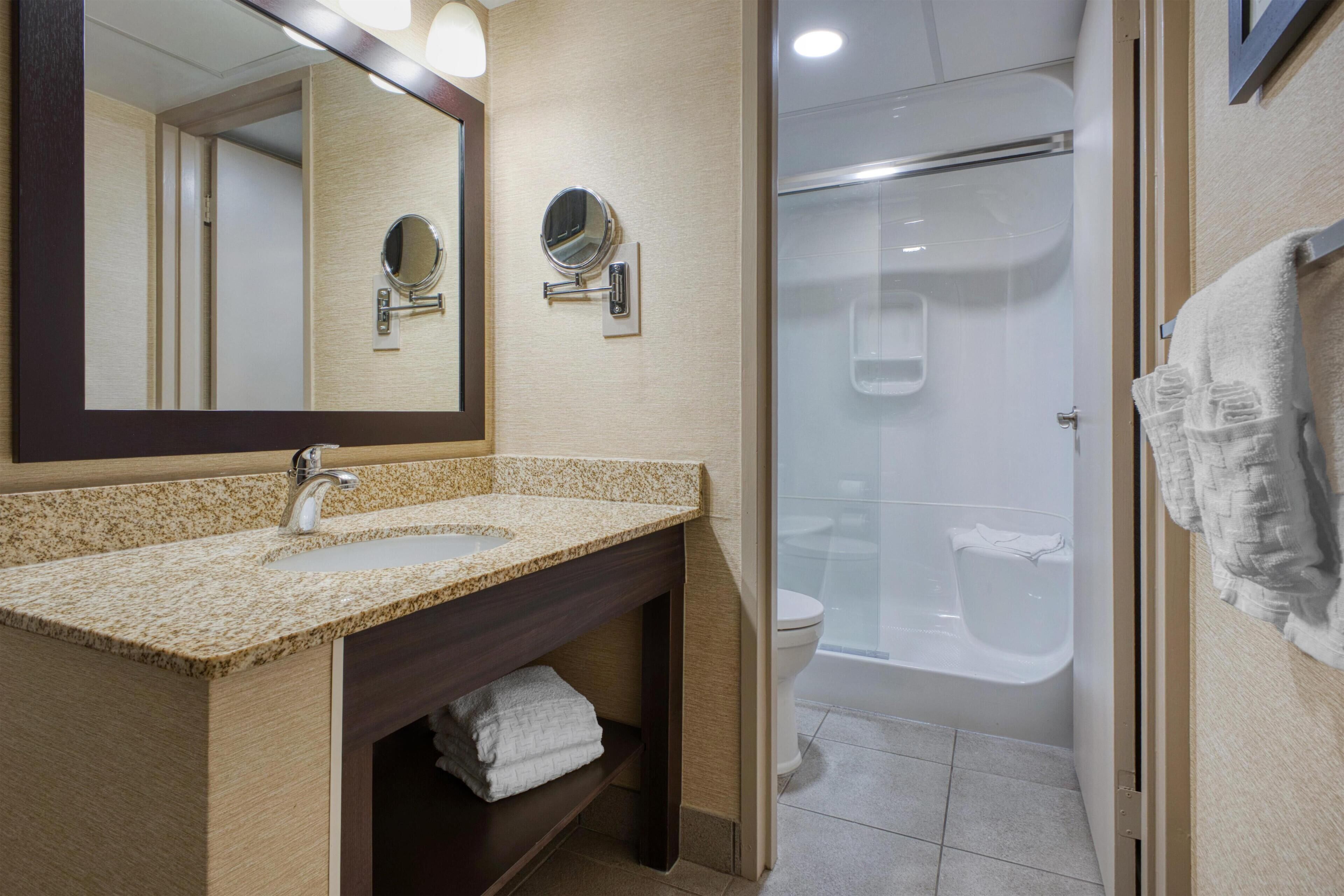 A bathroom with a granite countertop sink, a mirror, a towel rack, and a shower stall.