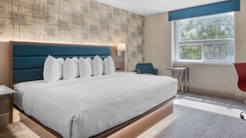 A modern hotel room with a large bed, a nightstand, a desk, a chair, a window with a view of trees, and a patterned wall.