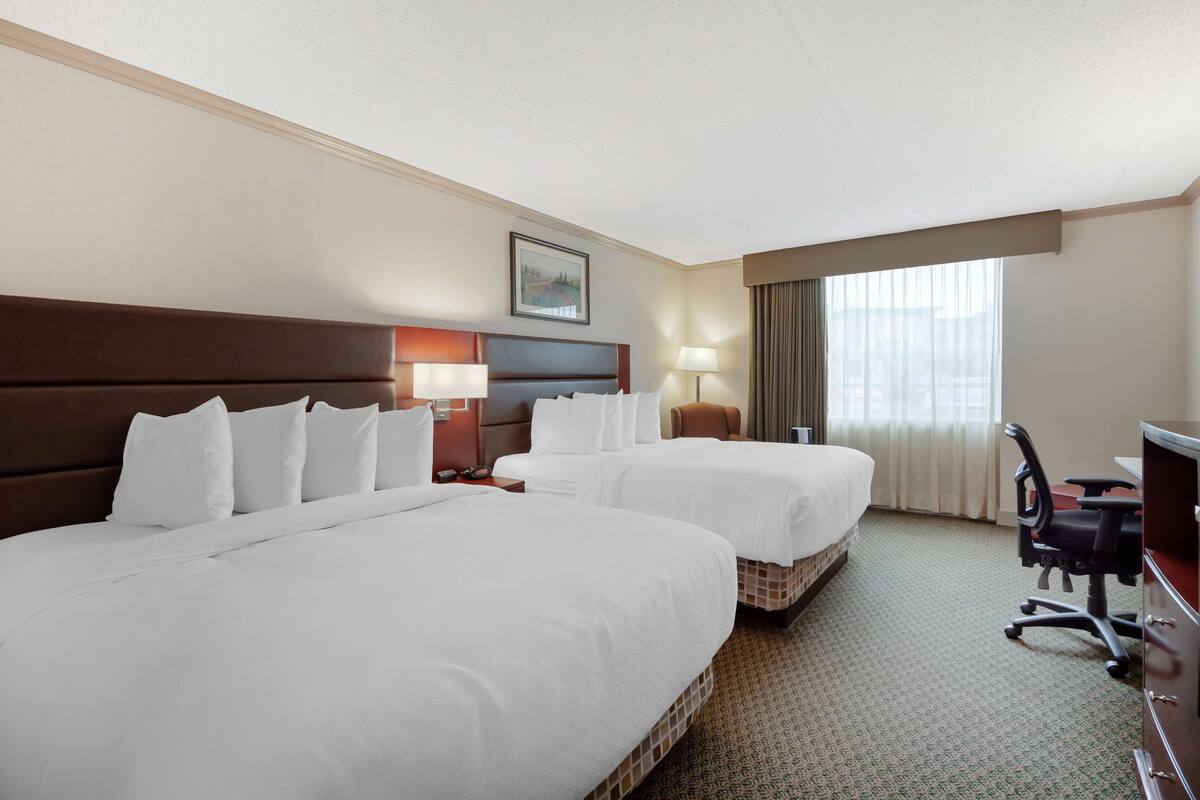 Habitación de hotel con dos camas grandes, un escritorio, una silla, una lámpara y un ventanal con cortinas.