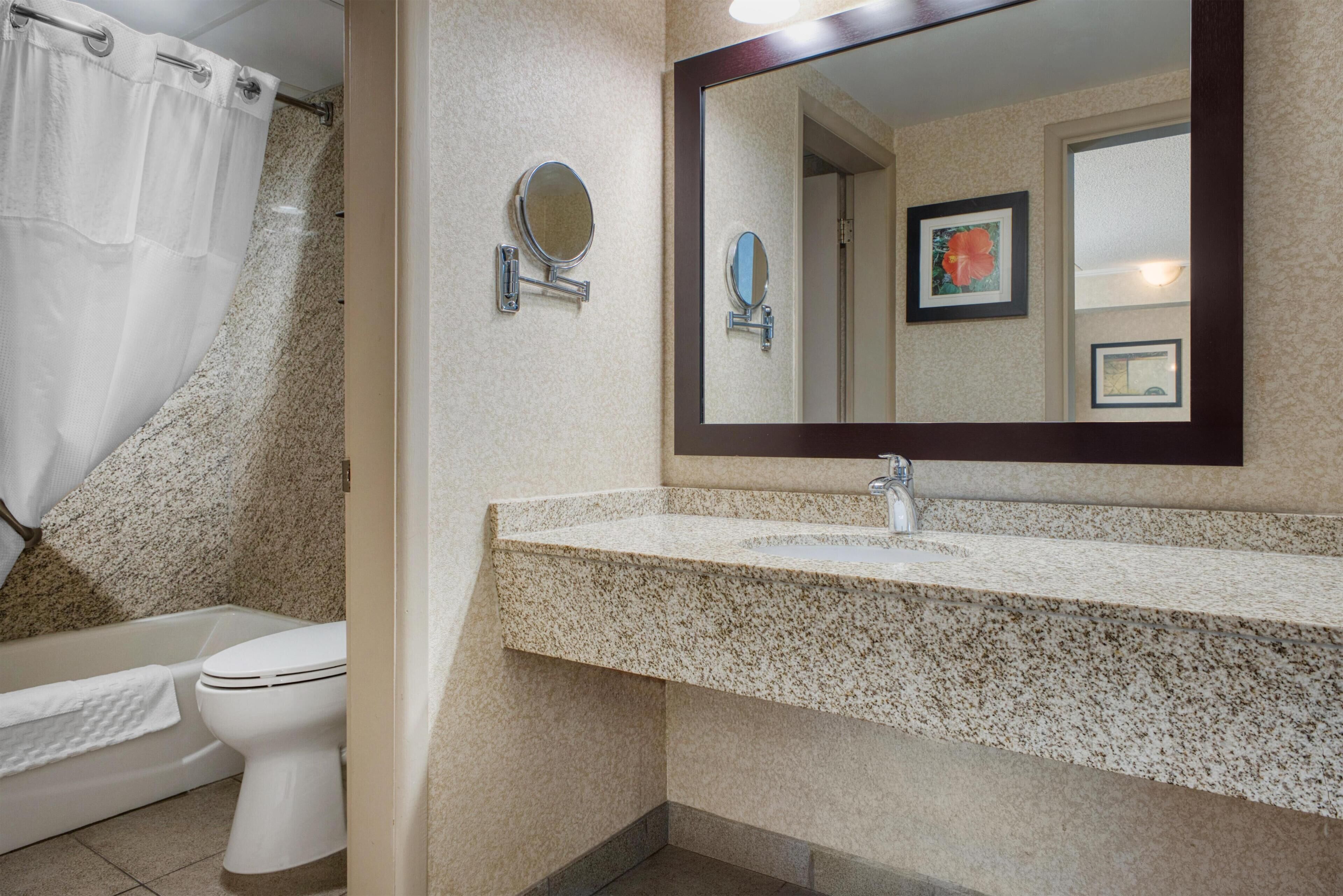 Une salle de bain avec des toilettes blanches, un lavabo doté d’un plan de travail en granit, un miroir et un rideau de douche.