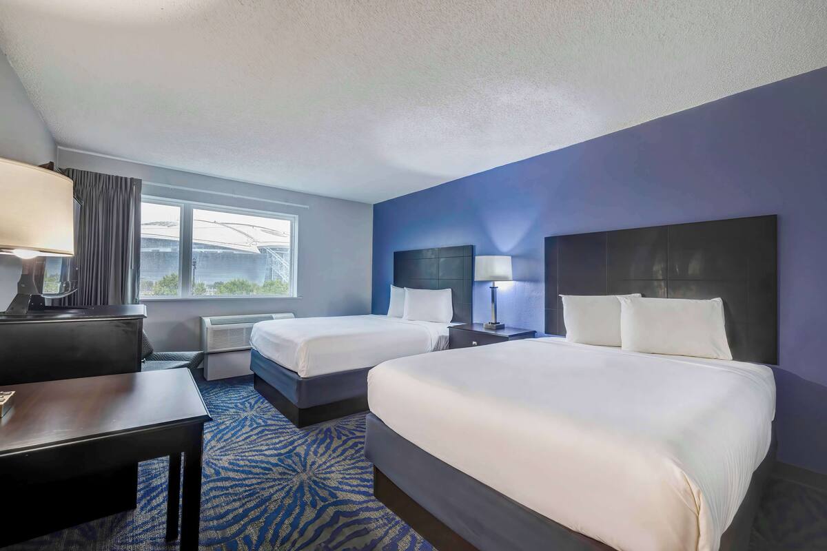A hotel room with two beds, a desk, a chair, a lamp, and a window with curtains.