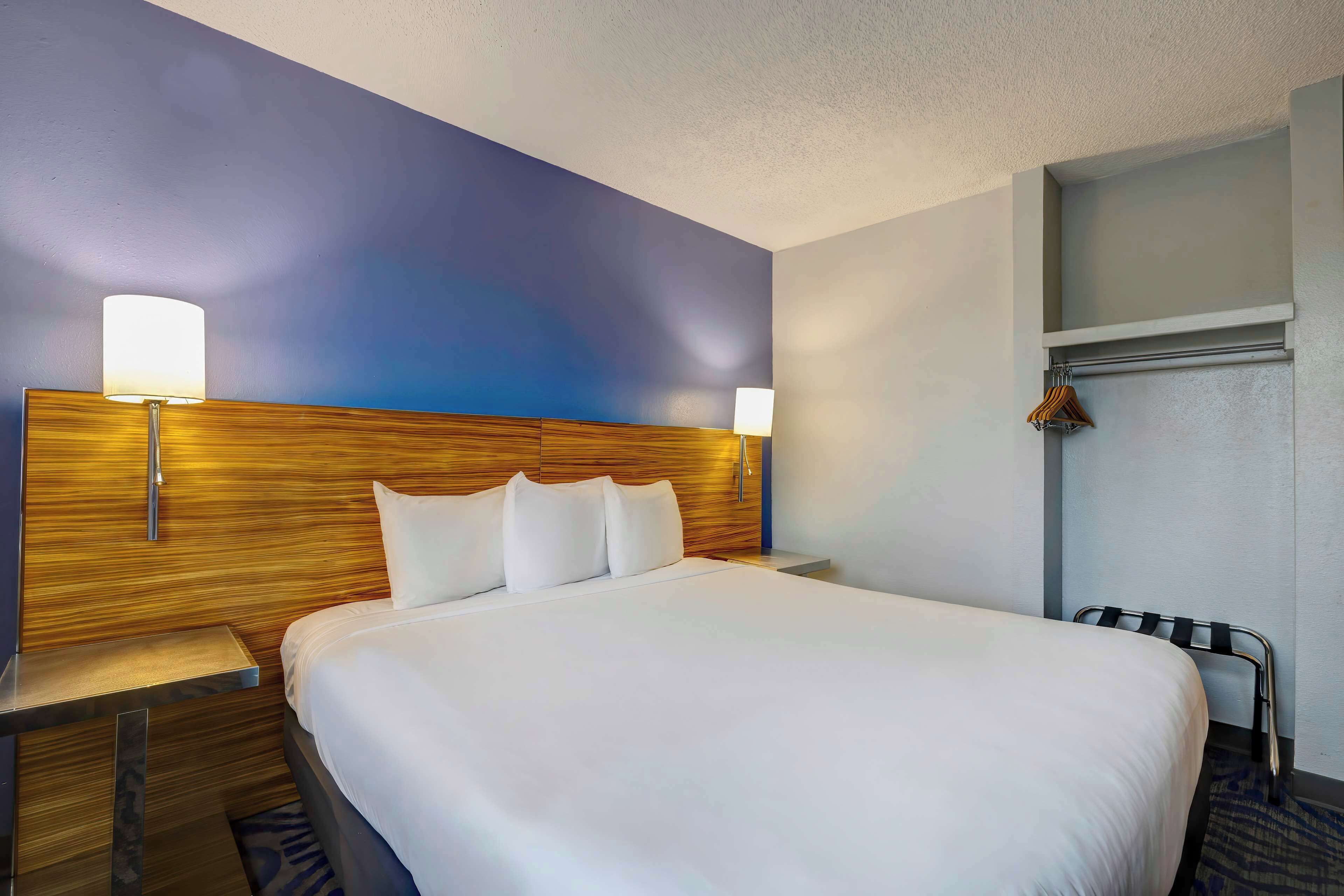 A hotel room with a large bed, wooden headboard, two bedside lamps, a nightstand, and a towel rack.