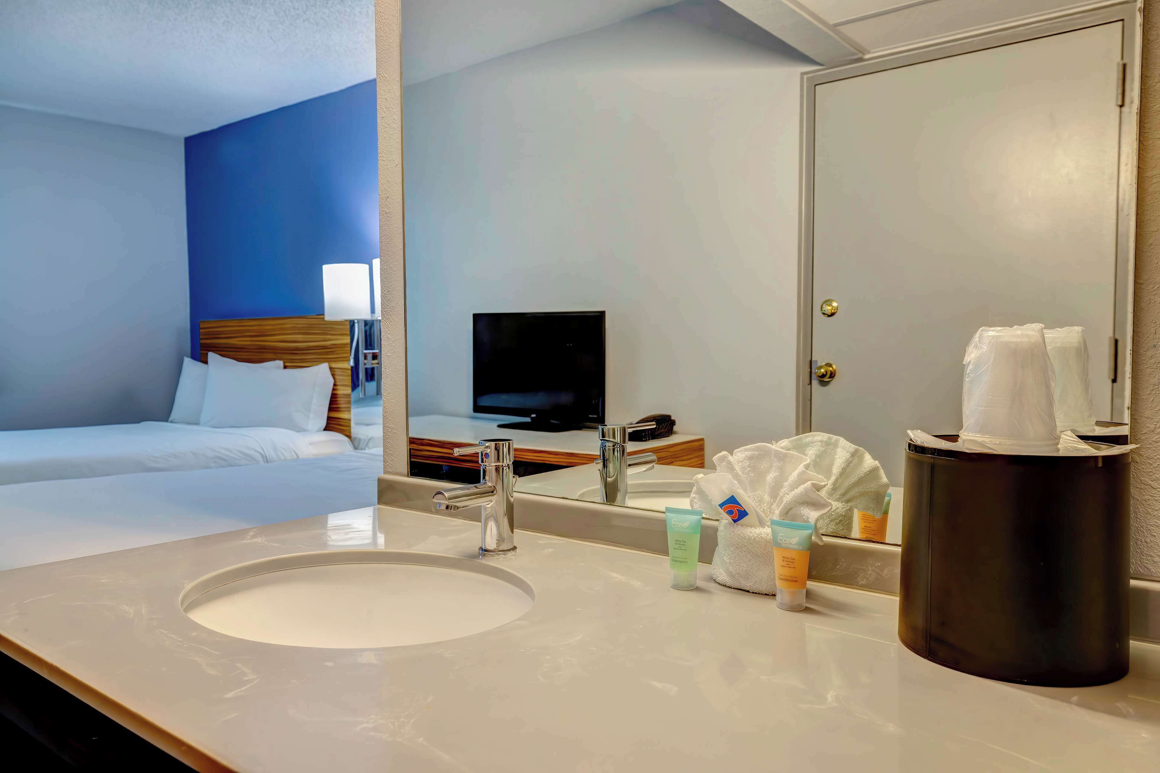 A hotel room with a bed, a large mirror, a sink, toiletries, and a television.