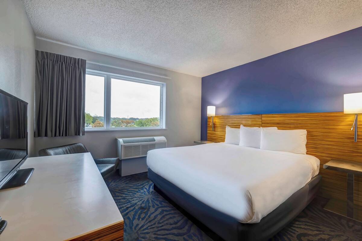 A hotel room with a large bed, a desk with a laptop, a window with curtains, a wall-mounted lamp, and a view of the outdoors.