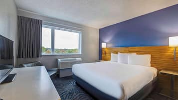 Ein Hotelzimmer mit einem großen Bett, einem Schreibtisch mit einem Laptop, einem Fenster mit Vorhängen, einer Wandlampe und Blick ins Freie.