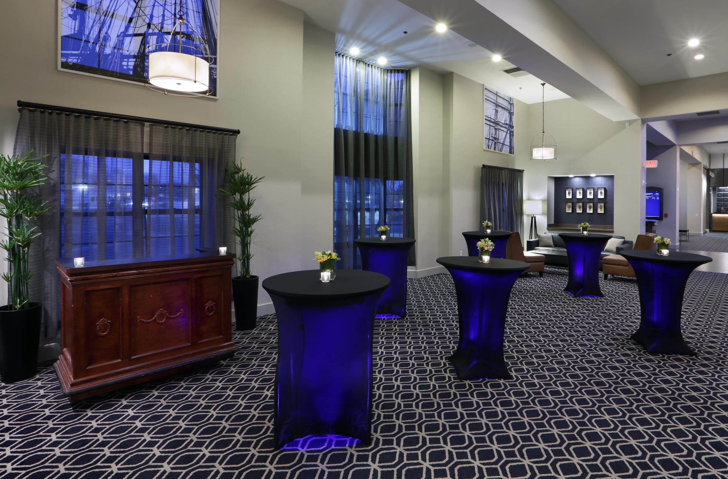 Een hotellobby met een modern design, met een bekleed vloerkleed, ronde tafels met blauwe verlichting en een groot houten kast.