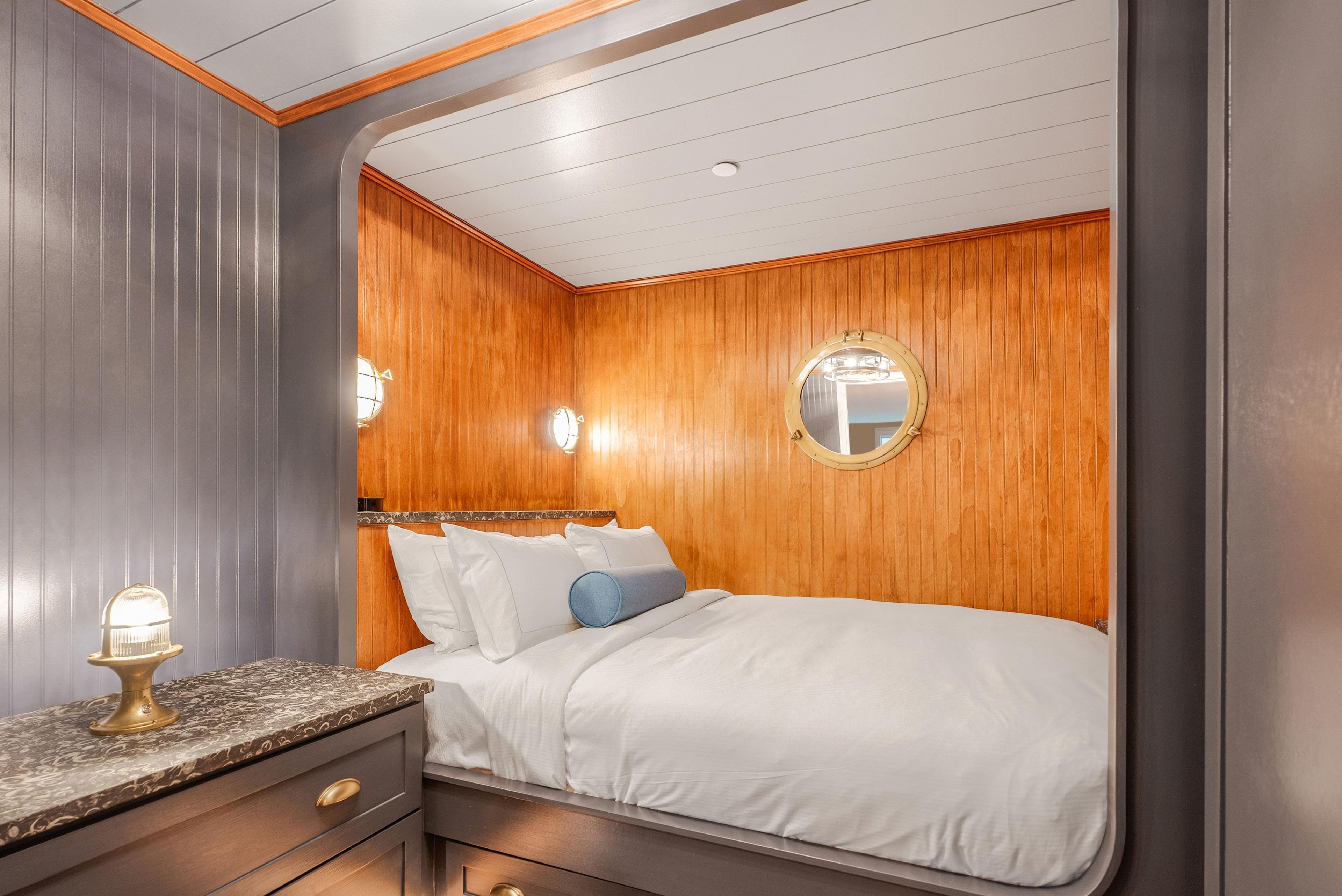 Un dormitorio con cabecera de madera, un espejo redondo, una cama con ropa blanca y una mesita de noche con lámpara.