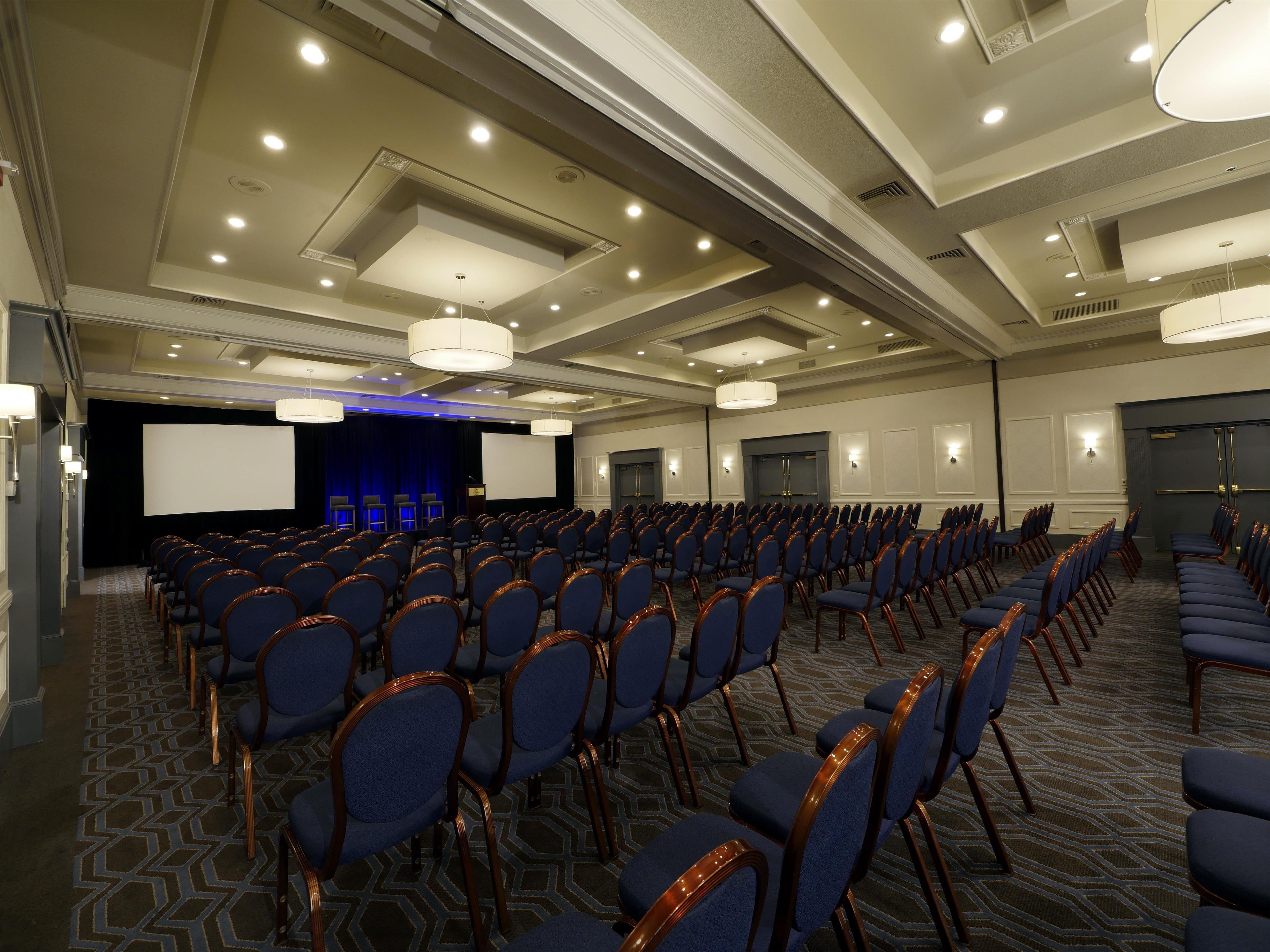 Un salón de conferencias con filas de sillas, una pantalla de proyector y un escenario.