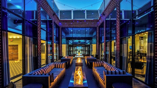 A modern outdoor lounge area with a long bench and cushioned seating, a central fire feature, and glass walls offering views of the interior. - Hilton Mystic (Mystic)