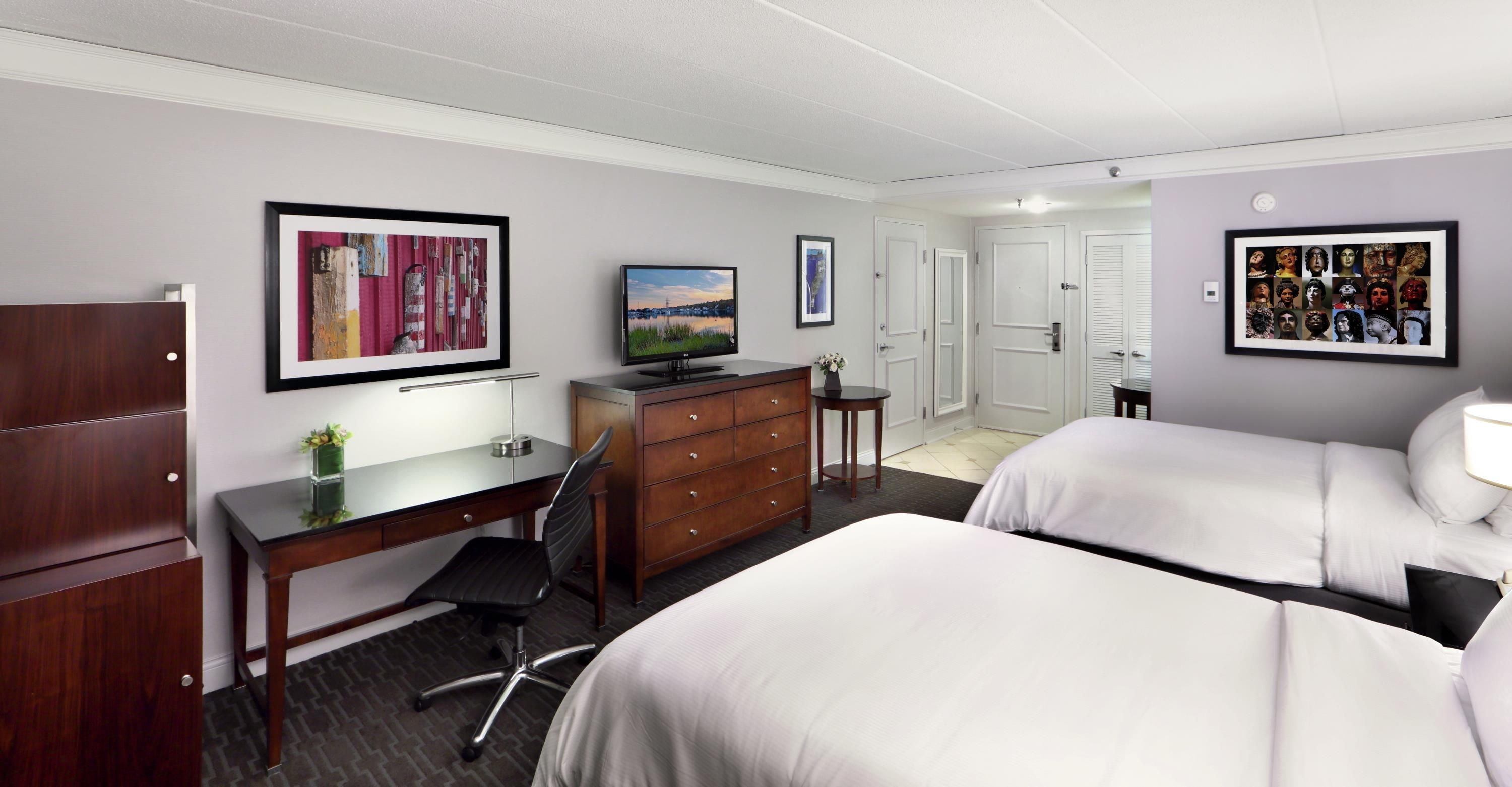 A hotel room with two beds, a desk, a chair, a dresser, a TV, and framed pictures on the walls.