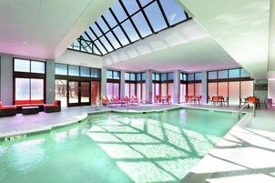 Una piscina interior con techos de vidrio transparente, zonas iluminadas por el sol con sillas de descanso y una estación de socorristas visible.