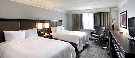 Quarto de hotel com duas camas, uma escrivaninha com cadeira, televisão e janela com cortinas.