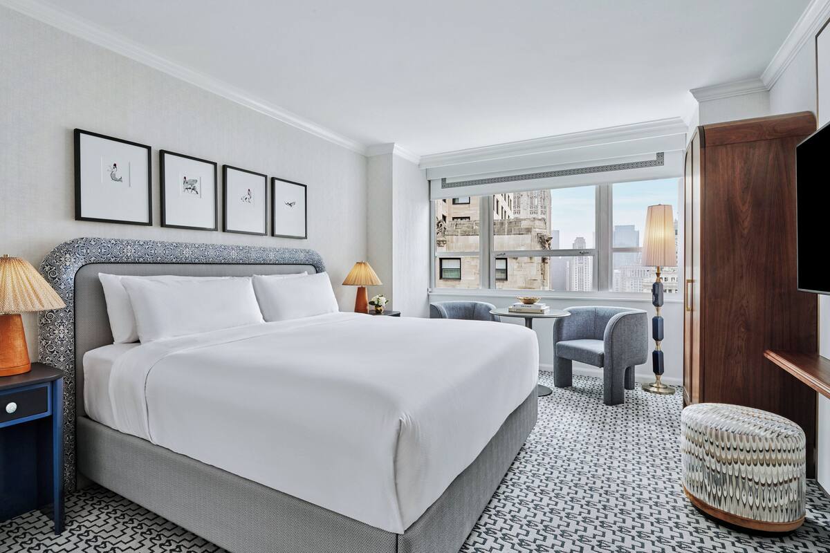 Habitación de hotel con una cama grande, un escritorio, una silla, un televisor y una ventana con vista a la ciudad.